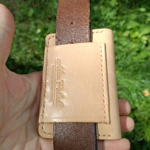 May include: A light tan leather belt loop accessory featuring a rectangular pocket with visible stitching. The leather is embossed with the text Alex Polak HANDMADE LEATHER GOODS. A dark brown leather belt is threaded through the vertical loop.