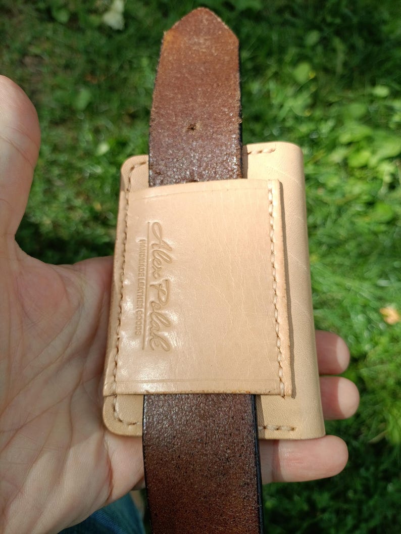 May include: A light tan leather belt loop accessory featuring a rectangular pocket with visible stitching. The leather is embossed with the text Alex Polak HANDMADE LEATHER GOODS. A dark brown leather belt is threaded through the vertical loop.