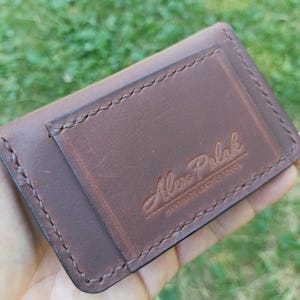Puede incluir: Un tarjetero de cuero marr&oacute;n oscuro con costuras visibles a lo largo de los bordes y un bolsillo rectangular en la parte delantera. El cuero est&aacute; grabado con el texto Alex Polak HANDMADE LEATHER GOODS en una combinaci&oacute;n de letra cursiva y bloque.