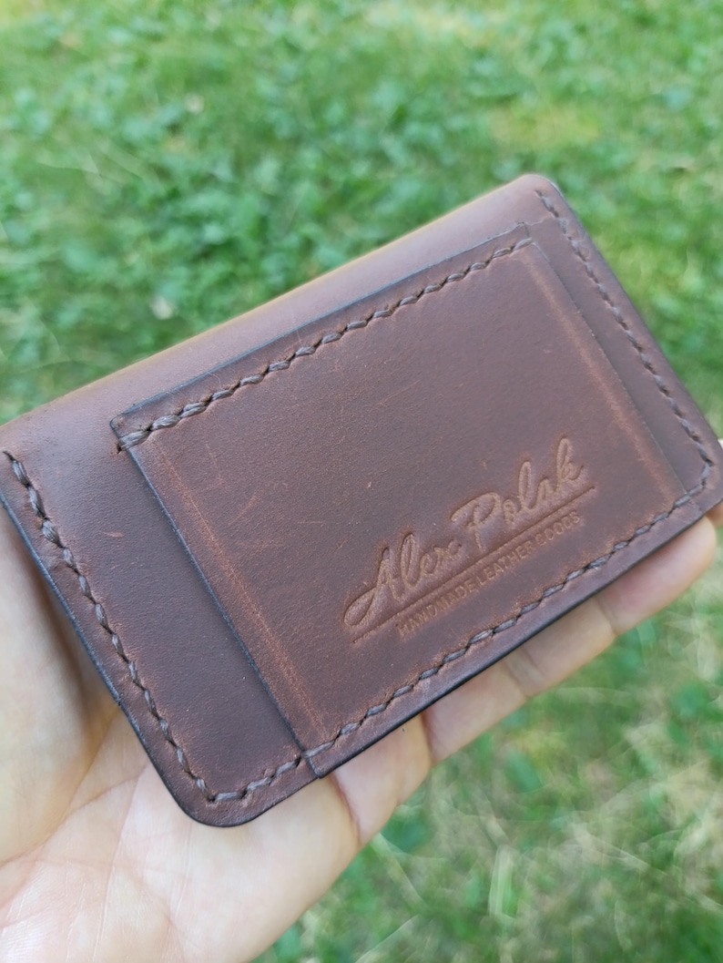 Puede incluir: Un tarjetero de cuero marr&oacute;n oscuro con costuras visibles a lo largo de los bordes y un bolsillo rectangular en la parte delantera. El cuero est&aacute; grabado con el texto Alex Polak HANDMADE LEATHER GOODS en una combinaci&oacute;n de letra cursiva y bloque.