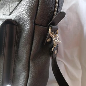 Puede incluir: Bolso mensajero de cuero negro con superficie texturizada. El bolso tiene una cremallera plateada, una correa de hombro desmontable y un bolsillo frontal. Tiene un asa en la parte superior y un cierre plateado para la correa.
