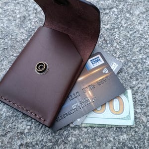 May include: A dark brown leather card holder with a brass snap button closure. The open flap reveals a grey bank card with the text EXIM BANK and a partial view of a one hundred dollar bill tucked inside the main compartment of the wallet.