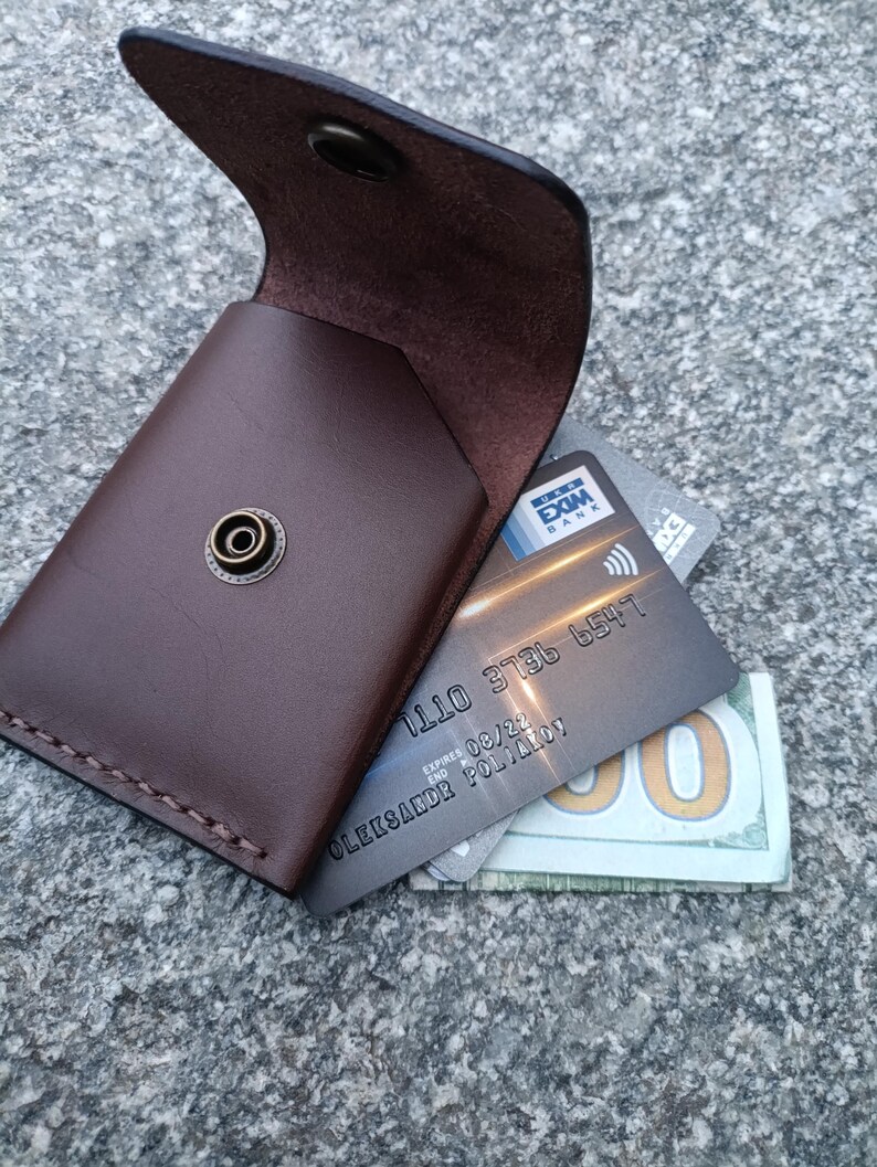 May include: A dark brown leather card holder with a brass snap button closure. The open flap reveals a grey bank card with the text EXIM BANK and a partial view of a one hundred dollar bill tucked inside the main compartment of the wallet.