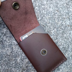 May include: A dark brown leather card holder with a flap closure secured by a metallic snap button. The interior features a smooth finish, while the exterior has a polished surface. A single rectangular card is partially visible inside the pocket.
