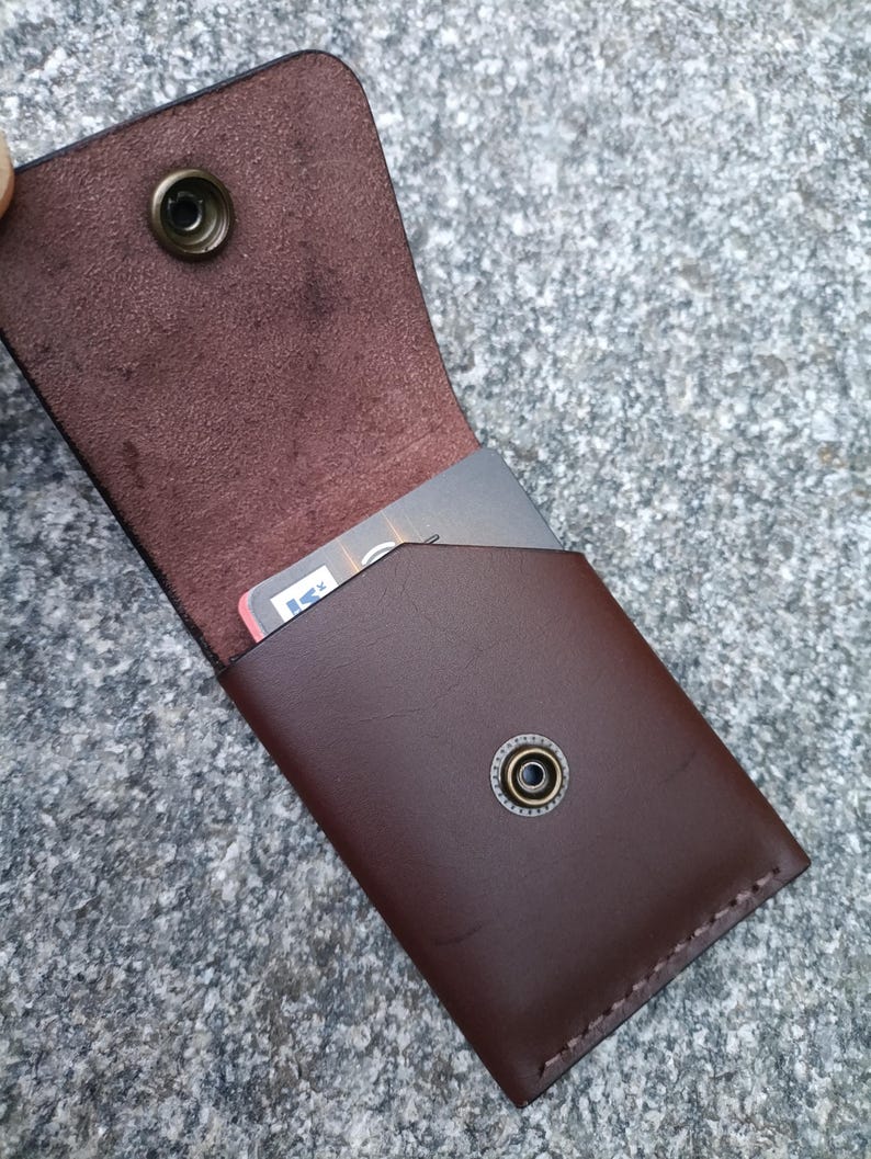 May include: A dark brown leather card holder with a flap closure secured by a metallic snap button. The interior features a smooth finish, while the exterior has a polished surface. A single rectangular card is partially visible inside the pocket.