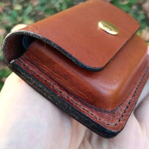 May include: A small, rectangular brown leather pouch featuring thick, dark-edged stitching along the perimeter and a single polished brass snap button closure on the front flap. The leather has a smooth, firm texture with visible grain.
