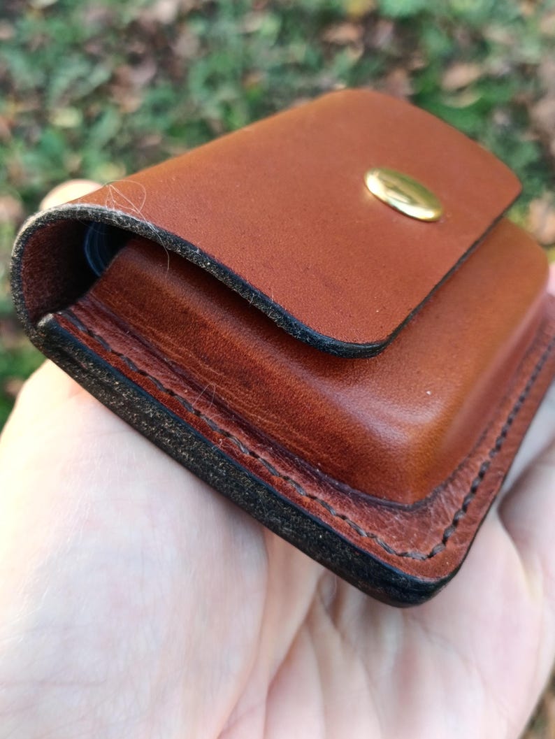May include: A small, rectangular brown leather pouch featuring thick, dark-edged stitching along the perimeter and a single polished brass snap button closure on the front flap. The leather has a smooth, firm texture with visible grain.