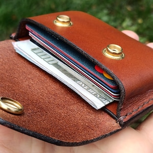 Puede incluir: Una cartera de cuero marr&oacute;n con exterior liso e interior texturizado. La cartera cuenta con dos broches de presi&oacute;n de metal dorado y contiene varias tarjetas de cr&eacute;dito apiladas y un billete de cien d&oacute;lares doblado en su interior.