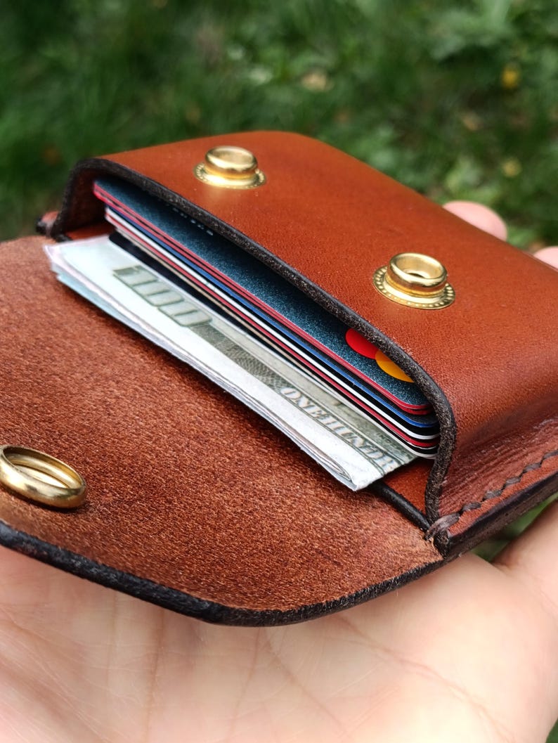 Puede incluir: Una cartera de cuero marr&oacute;n con exterior liso e interior texturizado. La cartera cuenta con dos broches de presi&oacute;n de metal dorado y contiene varias tarjetas de cr&eacute;dito apiladas y un billete de cien d&oacute;lares doblado en su interior.