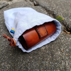K&ouml;nnte beinhalten: Ein hellblauer Kordelzugbeutel, der teilweise ge&ouml;ffnet ist und ein zylindrisches Lederetui in Braun zeigt. Das Lederetui hat einen gen&auml;hten Riemen. Der Beutel liegt auf einer Steinoberfl&auml;che. Ein kleines blau-gelbes Band ist am Beutel befestigt.