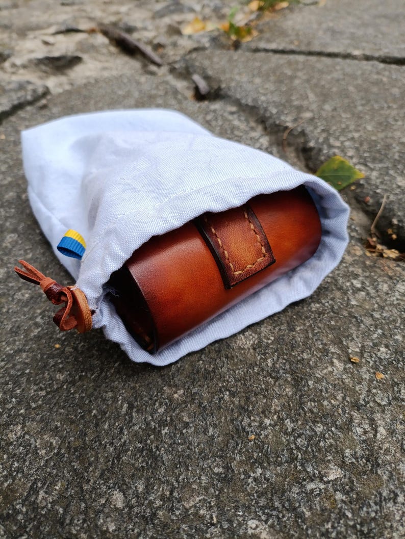 K&ouml;nnte beinhalten: Ein hellblauer Kordelzugbeutel, der teilweise ge&ouml;ffnet ist und ein zylindrisches Lederetui in Braun zeigt. Das Lederetui hat einen gen&auml;hten Riemen. Der Beutel liegt auf einer Steinoberfl&auml;che. Ein kleines blau-gelbes Band ist am Beutel befestigt.