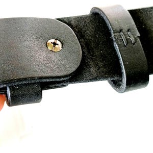 K&ouml;nnte beinhalten: Nahaufnahme eines schwarzen Lederg&uuml;rtels mit einer Metallschraube und gen&auml;hten Details. Der G&uuml;rtel hat eine Schnalle und eine Schlaufe. Das Leder wirkt strukturiert und abgenutzt, was auf Haltbarkeit und einen klassischen Stil hindeutet. Das Bild ist vor wei&szlig;em Hintergrund.