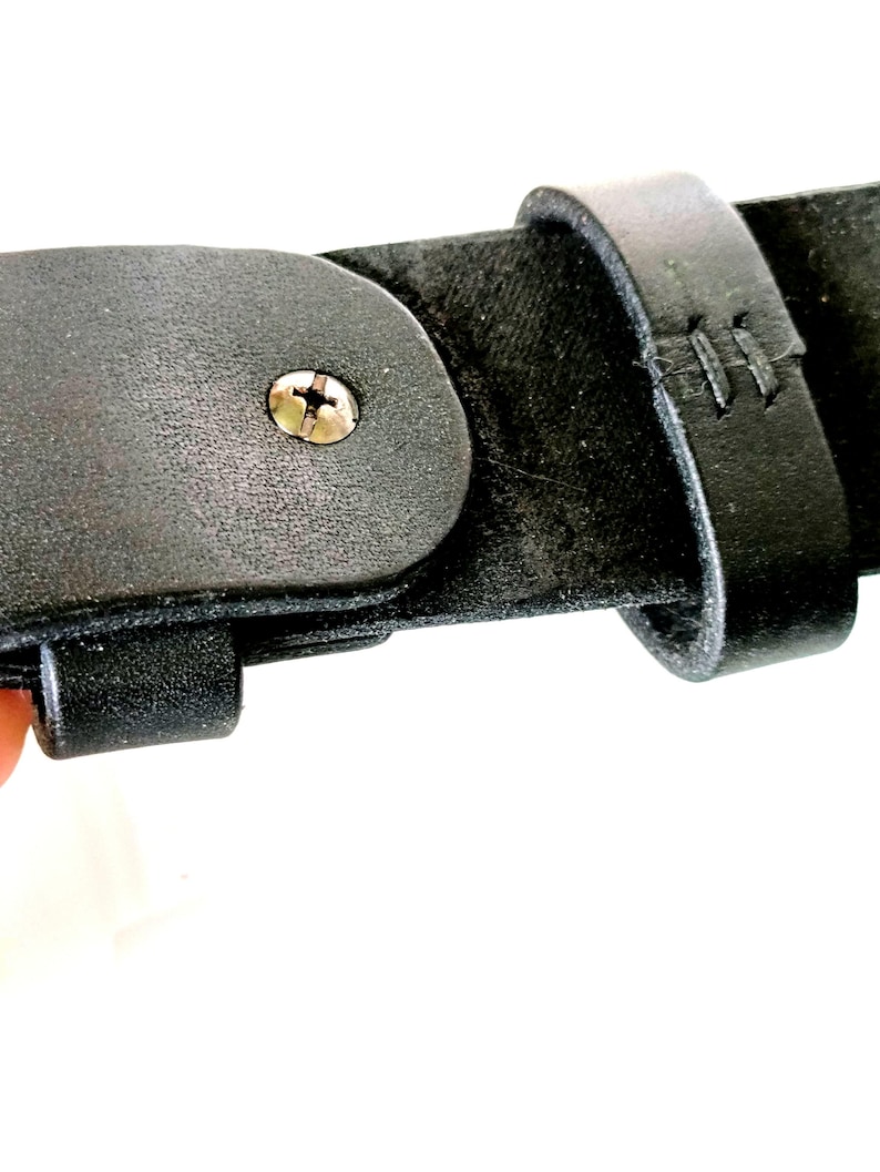 K&ouml;nnte beinhalten: Nahaufnahme eines schwarzen Lederg&uuml;rtels mit einer Metallschraube und gen&auml;hten Details. Der G&uuml;rtel hat eine Schnalle und eine Schlaufe. Das Leder wirkt strukturiert und abgenutzt, was auf Haltbarkeit und einen klassischen Stil hindeutet. Das Bild ist vor wei&szlig;em Hintergrund.
