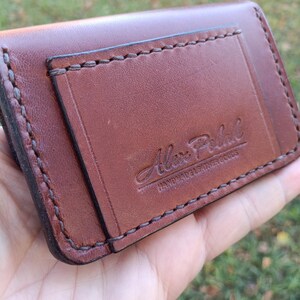 May include: A brown leather card holder featuring visible dark stitching along the edges and a rectangular pocket on the front. The leather is embossed with the text Alex Petal HANDMADE LEATHER GOODS in a script and block font style.