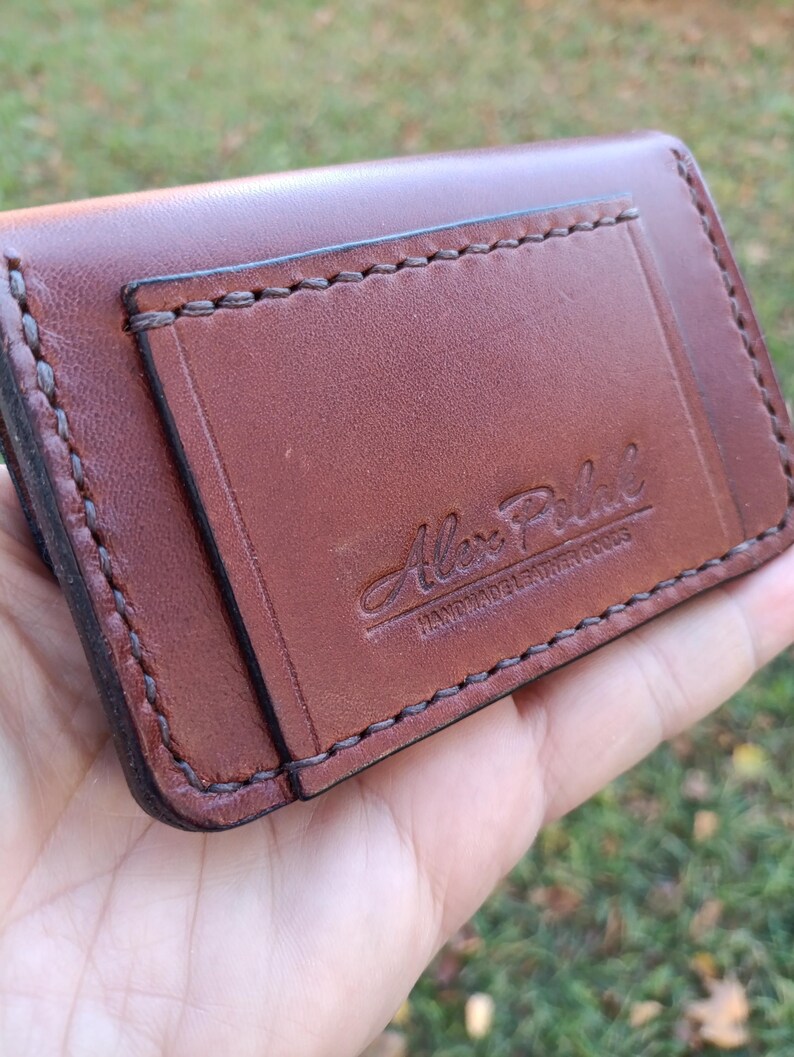 May include: A brown leather card holder featuring visible dark stitching along the edges and a rectangular pocket on the front. The leather is embossed with the text Alex Petal HANDMADE LEATHER GOODS in a script and block font style.