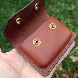 Puede incluir: Una bolsa de cuero marr&oacute;n con una solapa plegable que cuenta con dos broches de presi&oacute;n de lat&oacute;n y dos tachuelas de lat&oacute;n a juego en el cuerpo principal. Los bordes est&aacute;n cosidos con hilo oscuro y el cuero tiene un acabado pulido.