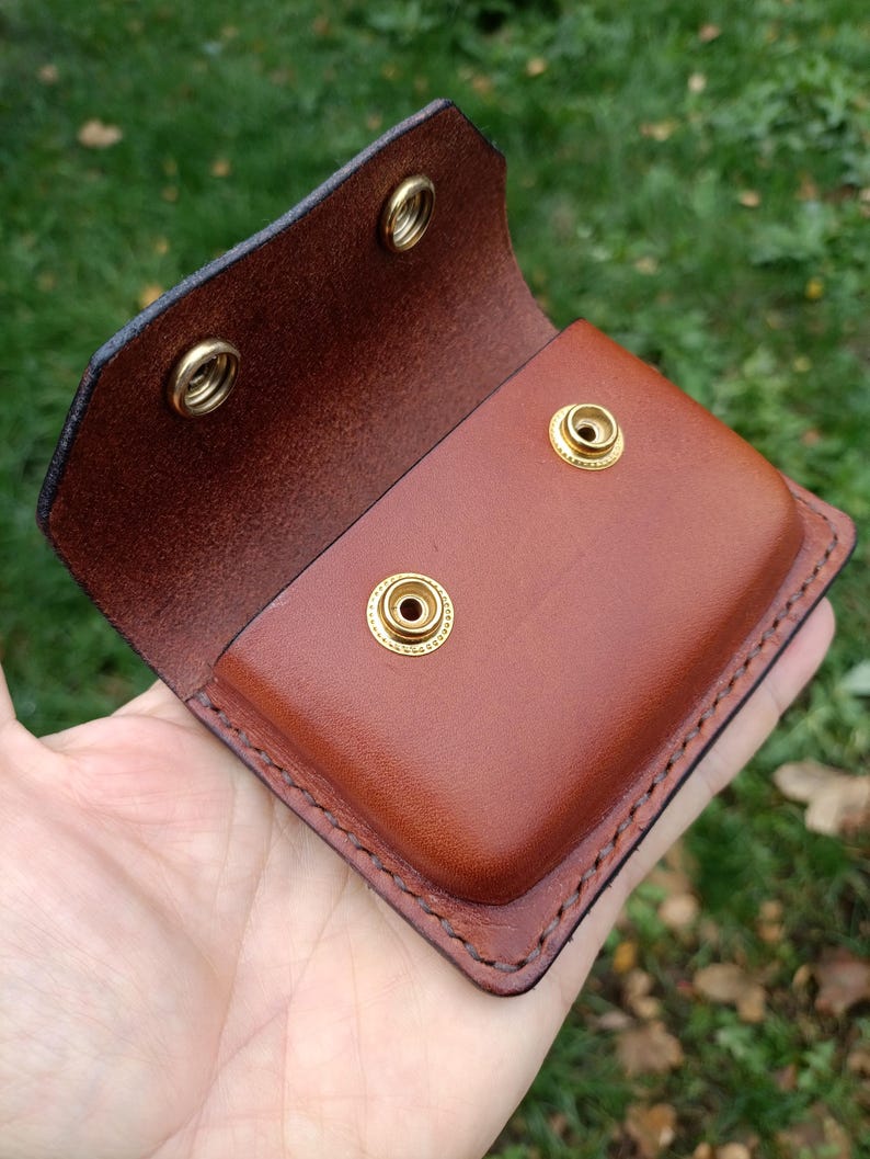 Puede incluir: Una bolsa de cuero marr&oacute;n con una solapa plegable que cuenta con dos broches de presi&oacute;n de lat&oacute;n y dos tachuelas de lat&oacute;n a juego en el cuerpo principal. Los bordes est&aacute;n cosidos con hilo oscuro y el cuero tiene un acabado pulido.