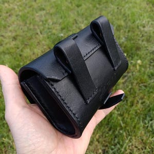 May include: A black leather pouch with a curved design, featuring two belt loops and a flap closure. The pouch is held in a hand against a backdrop of green grass. The stitching is visible along the edges.