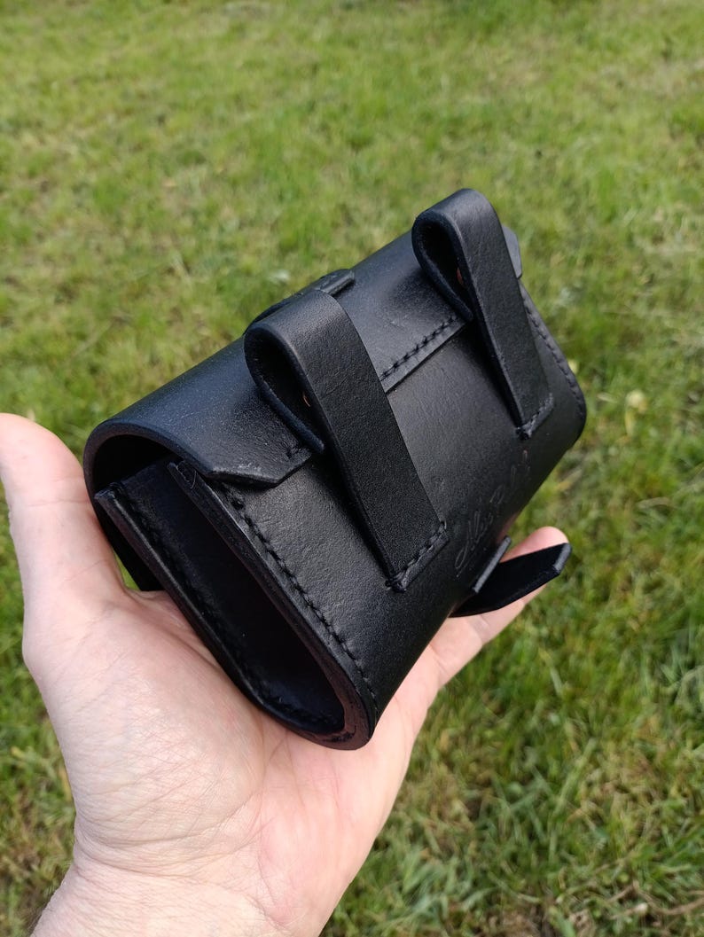 May include: A black leather pouch with a curved design, featuring two belt loops and a flap closure. The pouch is held in a hand against a backdrop of green grass. The stitching is visible along the edges.