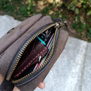Peut inclure: Une pochette en cuir marron fonc&eacute; avec une fermeture &eacute;clair en laiton. Le compartiment ouvert r&eacute;v&egrave;le un objet en cuir rouge fonc&eacute; avec des coutures visibles, un petit briquet Cricket turquoise et plusieurs cartes empil&eacute;es aux bords color&eacute;s.