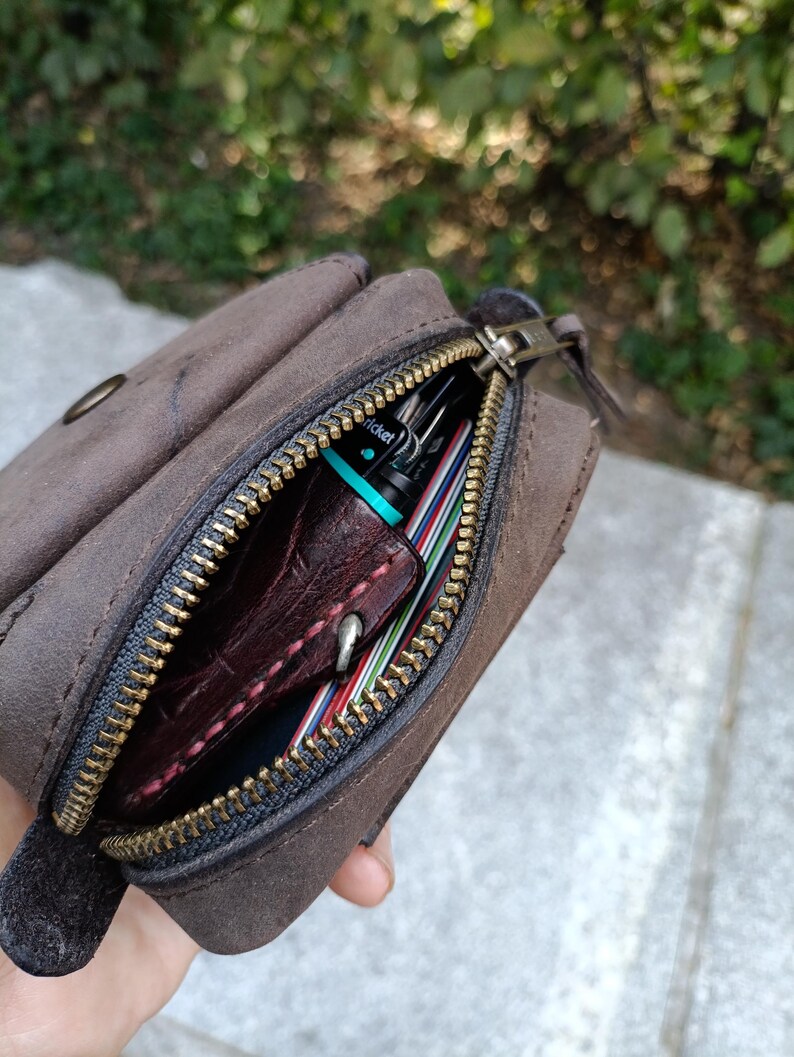 Peut inclure: Une pochette en cuir marron fonc&eacute; avec une fermeture &eacute;clair en laiton. Le compartiment ouvert r&eacute;v&egrave;le un objet en cuir rouge fonc&eacute; avec des coutures visibles, un petit briquet Cricket turquoise et plusieurs cartes empil&eacute;es aux bords color&eacute;s.