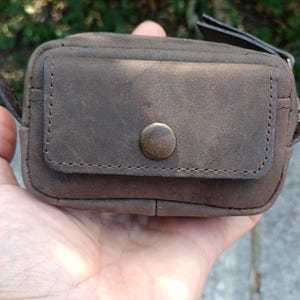 Peut inclure: Une petite pochette rectangulaire en cuir marron dot&eacute;e d'une poche frontale &agrave; rabat, ferm&eacute;e par un bouton-pression circulaire de couleur laiton. La pochette pr&eacute;sente des coutures visibles sur les bords du rabat et du corps, avec une finition mate.