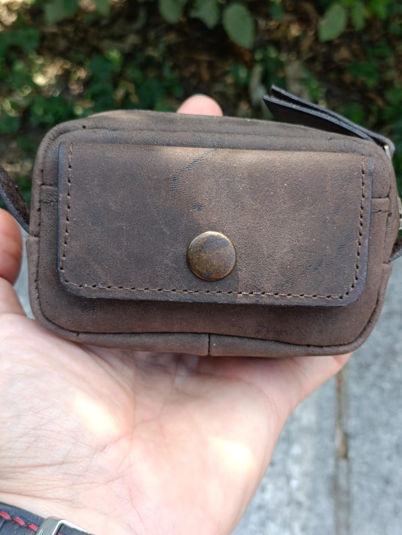 Peut inclure: Une petite pochette rectangulaire en cuir marron dot&eacute;e d'une poche frontale &agrave; rabat, ferm&eacute;e par un bouton-pression circulaire de couleur laiton. La pochette pr&eacute;sente des coutures visibles sur les bords du rabat et du corps, avec une finition mate.
