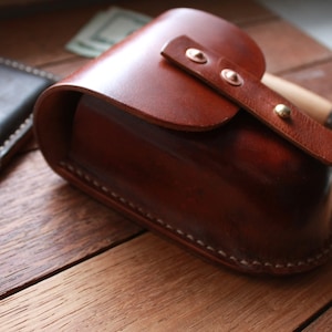 May include: A handcrafted, brown leather glasses case with a curved top and a strap secured by brass rivets. A dark leather wallet with a credit card is visible to the left. The case is on a wooden surface.