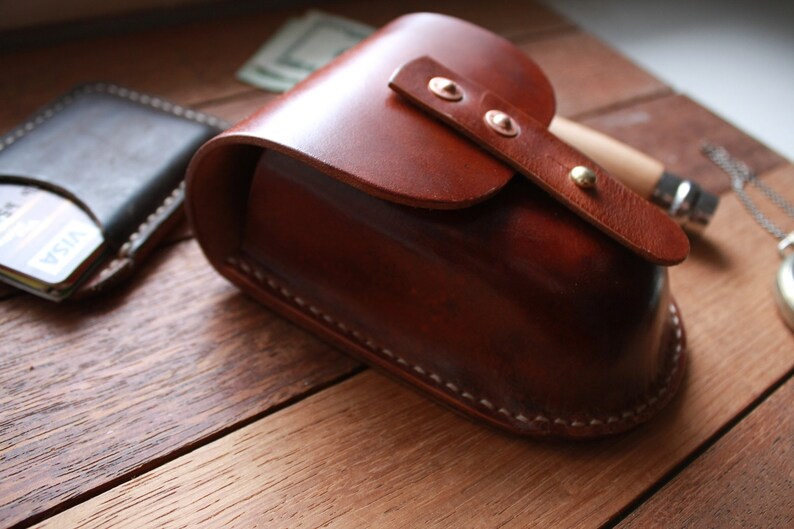 May include: A handcrafted, brown leather glasses case with a curved top and a strap secured by brass rivets. A dark leather wallet with a credit card is visible to the left. The case is on a wooden surface.