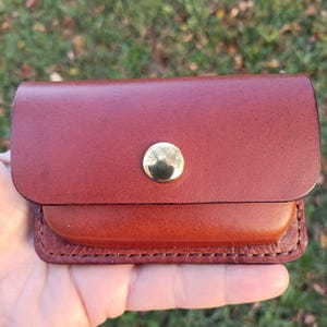 May include: A small rectangular brown leather pouch featuring a smooth flap closure secured by a single polished gold-tone metal snap button. The edges of the pouch are stitched with thick thread, showcasing a two-tone leather construction.