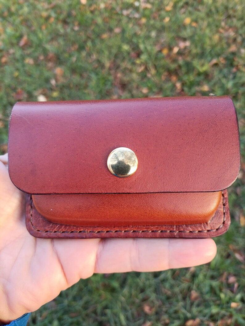 May include: A small rectangular brown leather pouch featuring a smooth flap closure secured by a single polished gold-tone metal snap button. The edges of the pouch are stitched with thick thread, showcasing a two-tone leather construction.