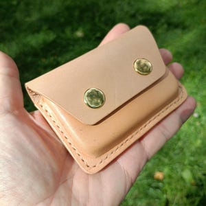 May include: A small, rectangular, light tan leather pouch featuring a fold-over flap secured by two polished gold-tone metal snap buttons. The edges of the pouch are joined with visible, thick, light-colored stitching along the perimeter.