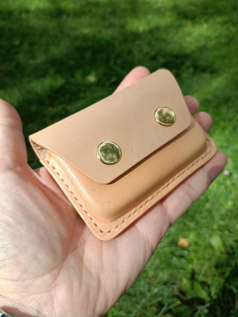 May include: A small, rectangular, light tan leather pouch featuring a fold-over flap secured by two polished gold-tone metal snap buttons. The edges of the pouch are joined with visible, thick, light-colored stitching along the perimeter.