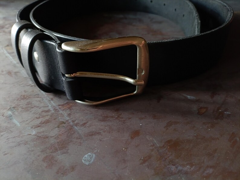 Puede incluir: Un cintur&oacute;n de cuero negro con un acabado liso y mate, que presenta una hebilla de metal en tono dorado pulido. El cintur&oacute;n incluye una trabilla de cuero a juego y un remache met&aacute;lico que asegura la correa cerca de la hebilla.