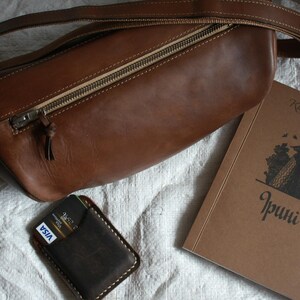 May include: A brown leather shoulder bag with a zipper closure and a credit card holder with a VISA logo. The bag is lying on a white surface next to a brown book with the text "Kisa Celebiyanli" and "Irinci" on the cover.