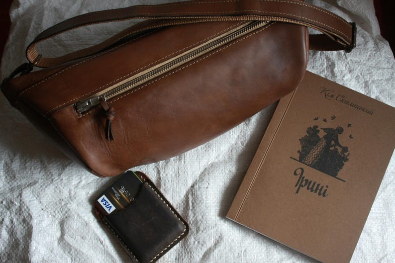 May include: A brown leather shoulder bag with a zipper closure and a credit card holder with a VISA logo. The bag is lying on a white surface next to a brown book with the text "Kisa Celebiyanli" and "Irinci" on the cover.