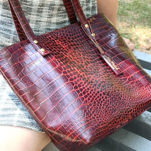 May include: A deep red tote bag with a glossy crocodile skin texture. The bag features two shoulder straps attached with gold-toned metal rivets and a small rectangular leather tag hanging from one strap with the embossed text Alex Pollak.