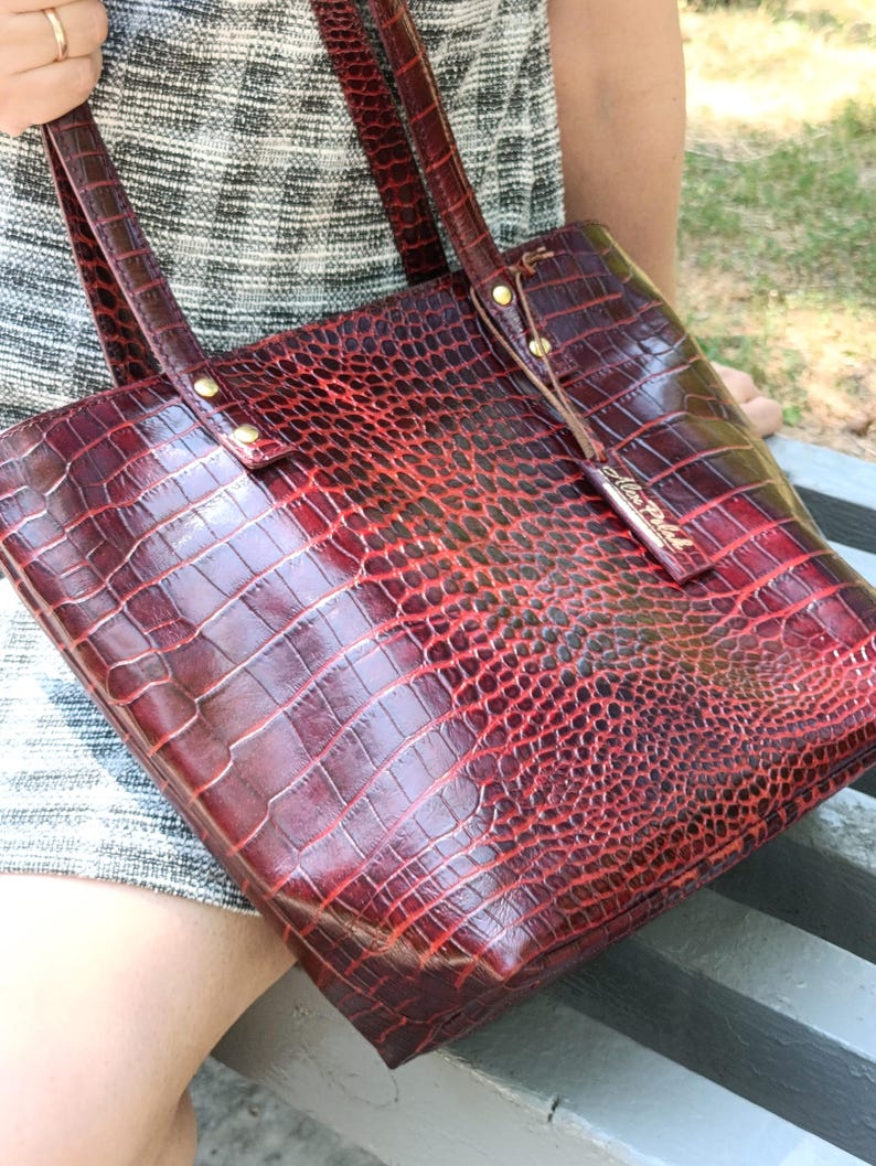 May include: A deep red tote bag with a glossy crocodile skin texture. The bag features two shoulder straps attached with gold-toned metal rivets and a small rectangular leather tag hanging from one strap with the embossed text Alex Pollak.