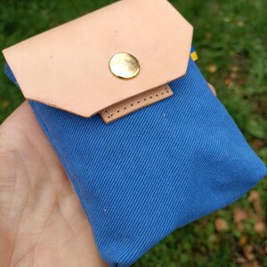 Puede incluir: Una peque&ntilde;a bolsa de lona azul con una solapa de cuero color canela claro, asegurada con un solo bot&oacute;n de presi&oacute;n de metal dorado pulido. La bolsa rectangular tiene una textura de tejido diagonal y un dise&ntilde;o sencillo.