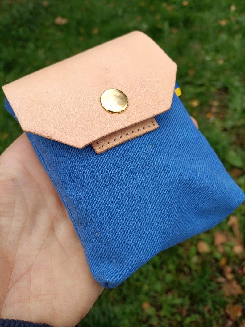 Puede incluir: Una peque&ntilde;a bolsa de lona azul con una solapa de cuero color canela claro, asegurada con un solo bot&oacute;n de presi&oacute;n de metal dorado pulido. La bolsa rectangular tiene una textura de tejido diagonal y un dise&ntilde;o sencillo.