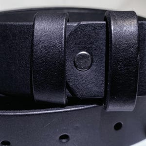 May include: Close-up of a black leather belt with a silver buckle. The belt has a classic design with a simple, elegant aesthetic. The leather appears smooth and durable, with visible stitching and a metal buckle.