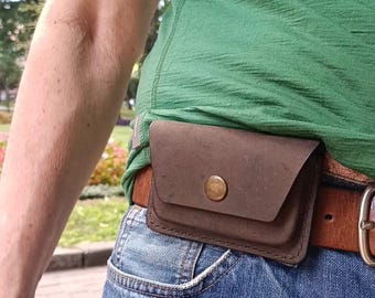 Könnte beinhalten: Eine dunkelbraune Gürteltasche aus Leder mit einem einzelnen messingfarbenen Druckknopfverschluss. Die rechteckige Tasche ist an einem hellbraunen Ledergürtel befestigt, der über einer blauen Jeans und einem grünen T-Shirt getragen wird.