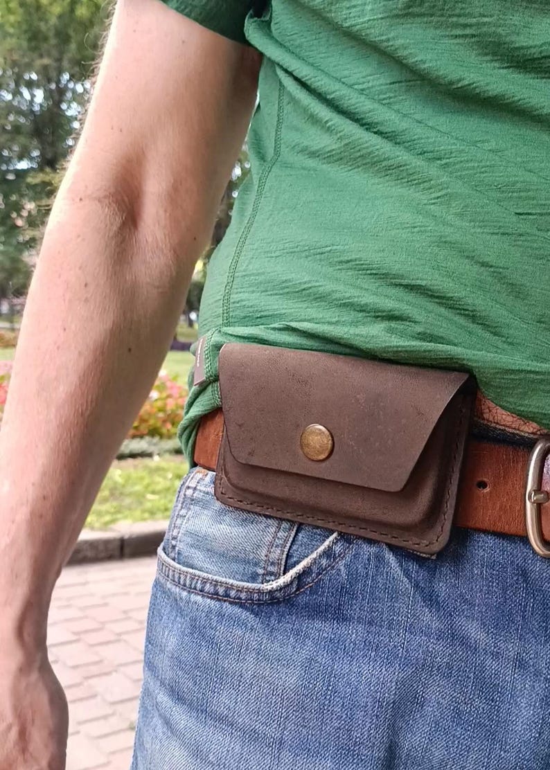 May include: A dark brown leather belt pouch with a single brass-colored snap button closure. The rectangular pouch is attached to a tan leather belt worn over blue denim jeans and a green t-shirt, providing a compact storage solution.
