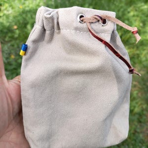 May include: A beige drawstring pouch made of a textured fabric with a silver metal grommet at the top. A thin, dark brown leather cord is threaded through the grommet. A small, rectangular tag with blue and yellow horizontal stripes is attached to the side.