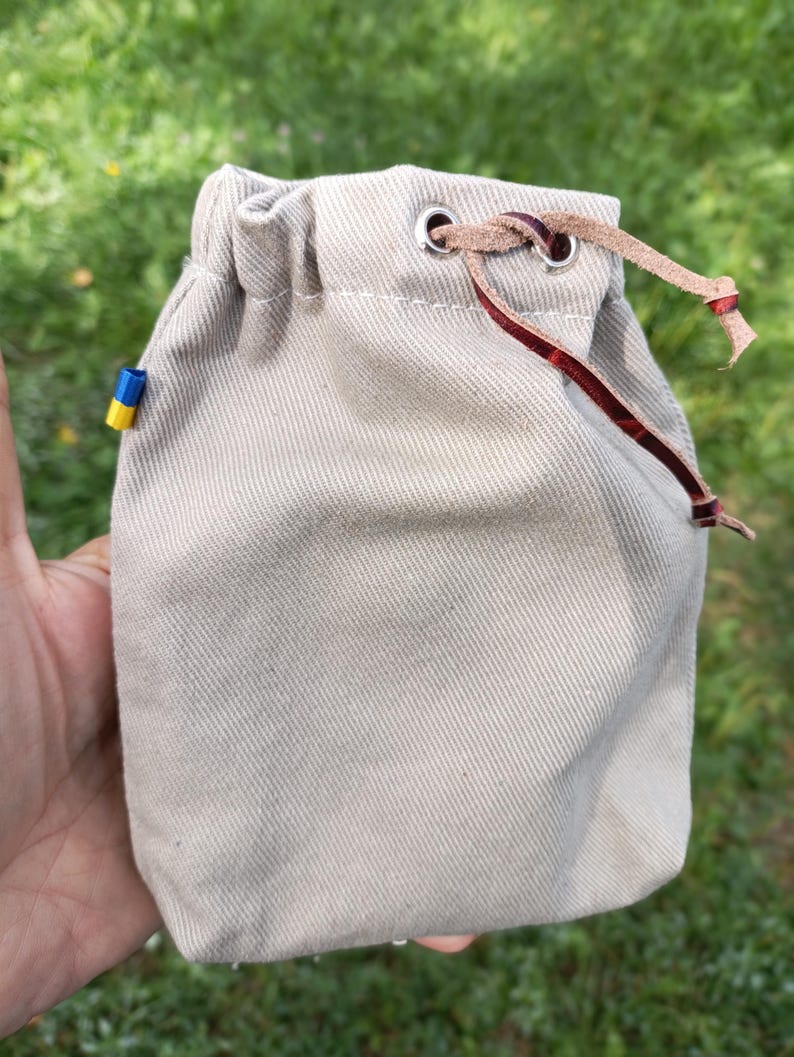 May include: A beige drawstring pouch made of a textured fabric with a silver metal grommet at the top. A thin, dark brown leather cord is threaded through the grommet. A small, rectangular tag with blue and yellow horizontal stripes is attached to the side.