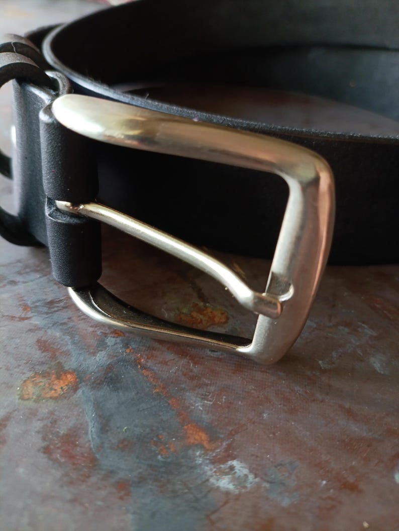 Puede incluir: Un cintur&oacute;n negro con una correa de acabado mate y liso, junto a una hebilla de metal plateado pulido. La hebilla presenta un marco rectangular con esquinas redondeadas y un perno, sujeta a la correa mediante una trabilla negra a juego.