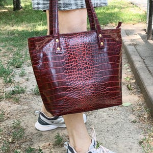 May include: A deep red tote bag with a glossy crocodile skin texture, featuring two vertical shoulder straps attached with gold-toned metal rivets. The bag has a structured rectangular shape and a smooth, shiny finish across the entire surface.