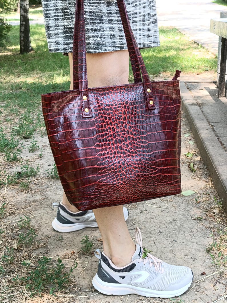 May include: A deep red tote bag with a glossy crocodile skin texture, featuring two vertical shoulder straps attached with gold-toned metal rivets. The bag has a structured rectangular shape and a smooth, shiny finish across the entire surface.