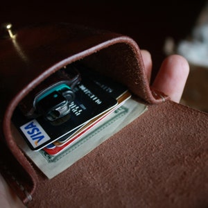 May include: A brown leather wallet with a flap that opens to reveal credit cards and cash. The wallet has a brass button closure.