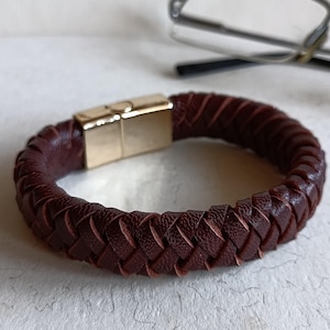 May include: A brown leather bracelet with a gold magnetic clasp. The bracelet is braided and has a simple design.